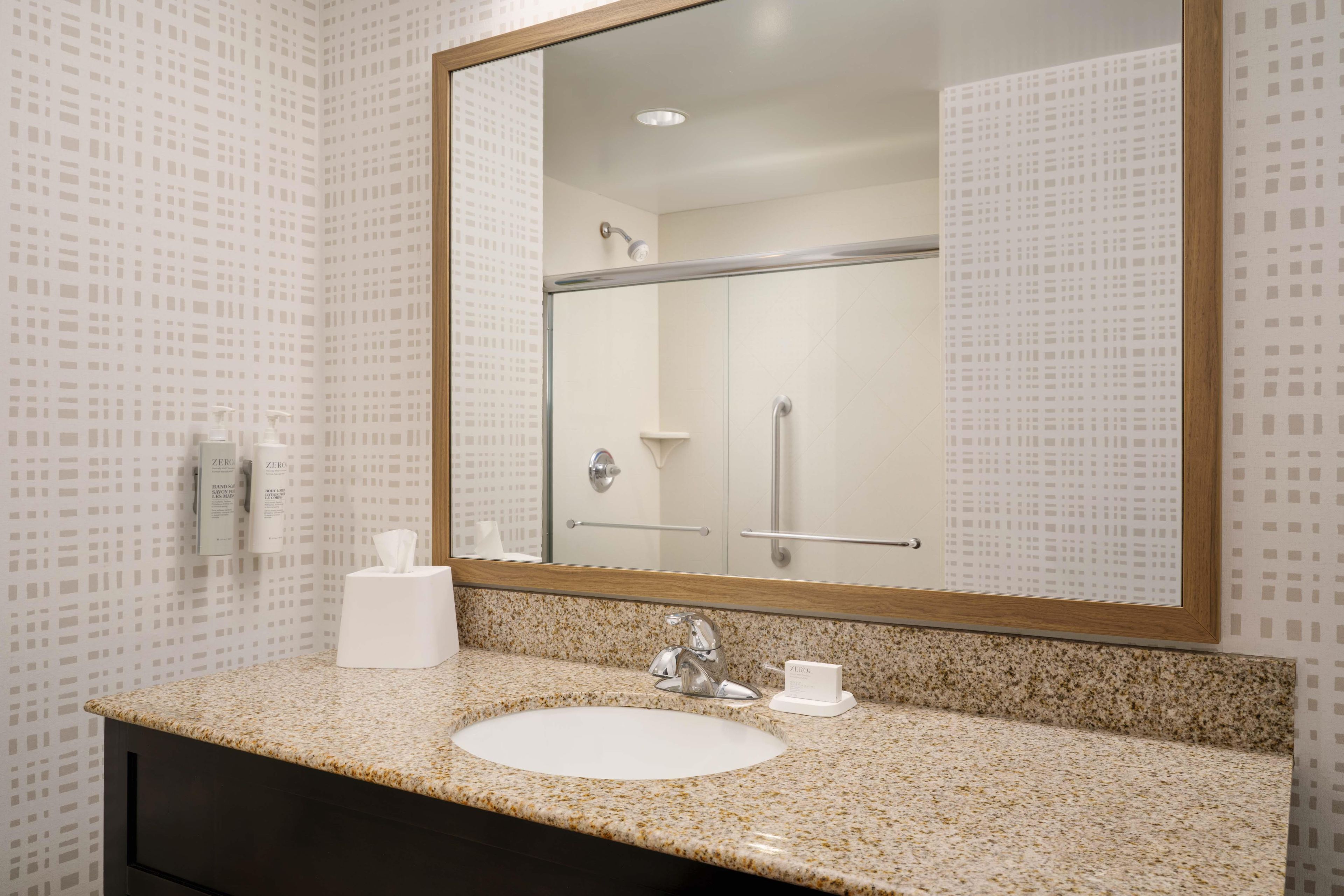A bathroom with a large mirror, a sink, a towel dispenser, and a shower.