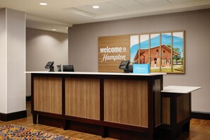 Un mostrador de recepción con un cartel que dice 'Welcome to Hampton', un teléfono y un cartel de 'Hilton'.