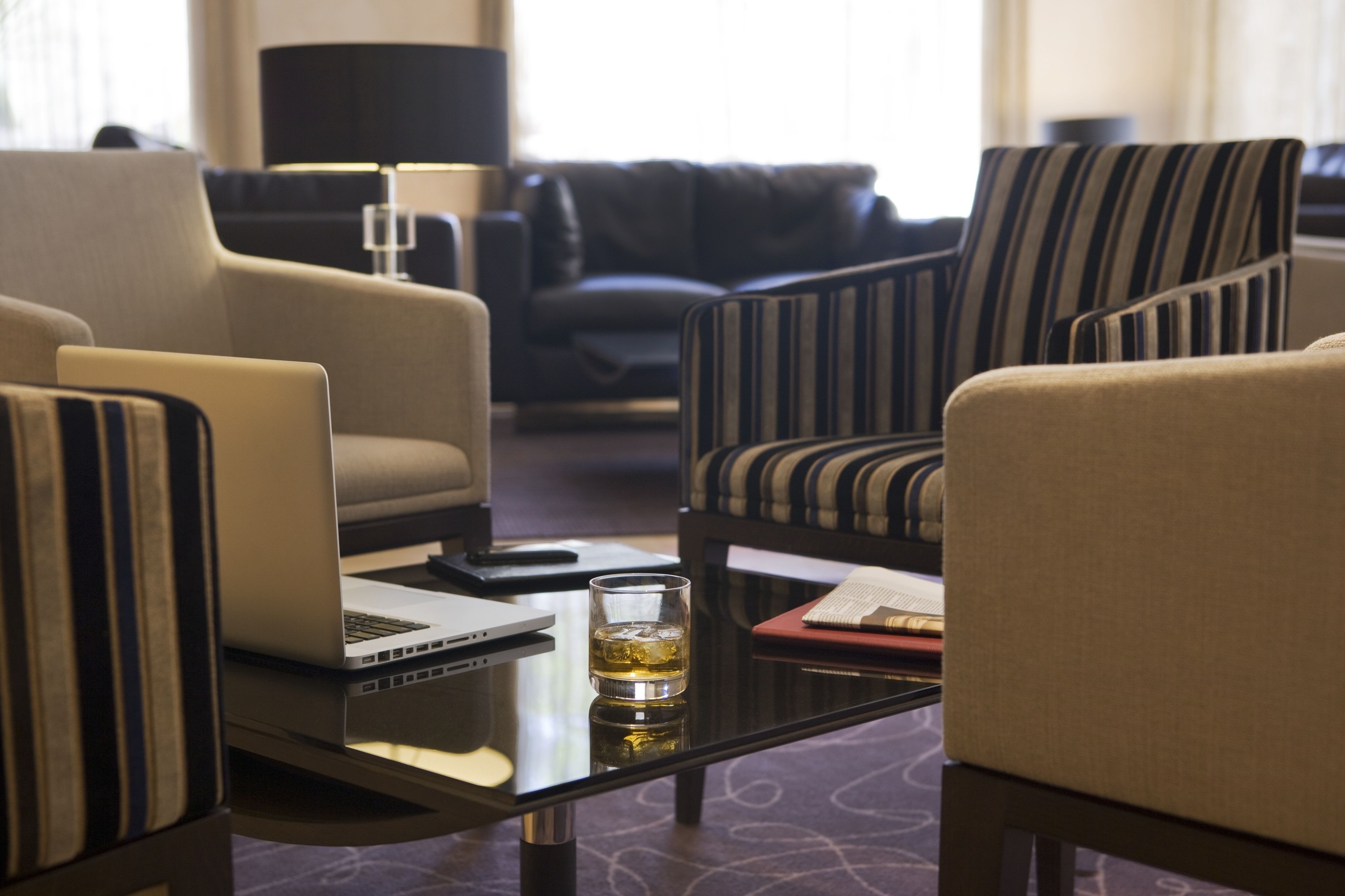 Hotelkamer met een bank, gestreepte fauteuils, een salontafel met een laptop en een whiskeyglas, en een lamp op de achtergrond.