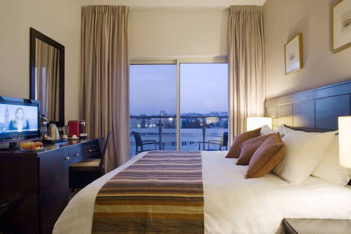 A hotel room with a large bed, a desk with a television, and a view of a cityscape through the window.