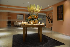 A lobby with a round table featuring a floral arrangement and candles, a framed painting on the wall, and a large potted plant.