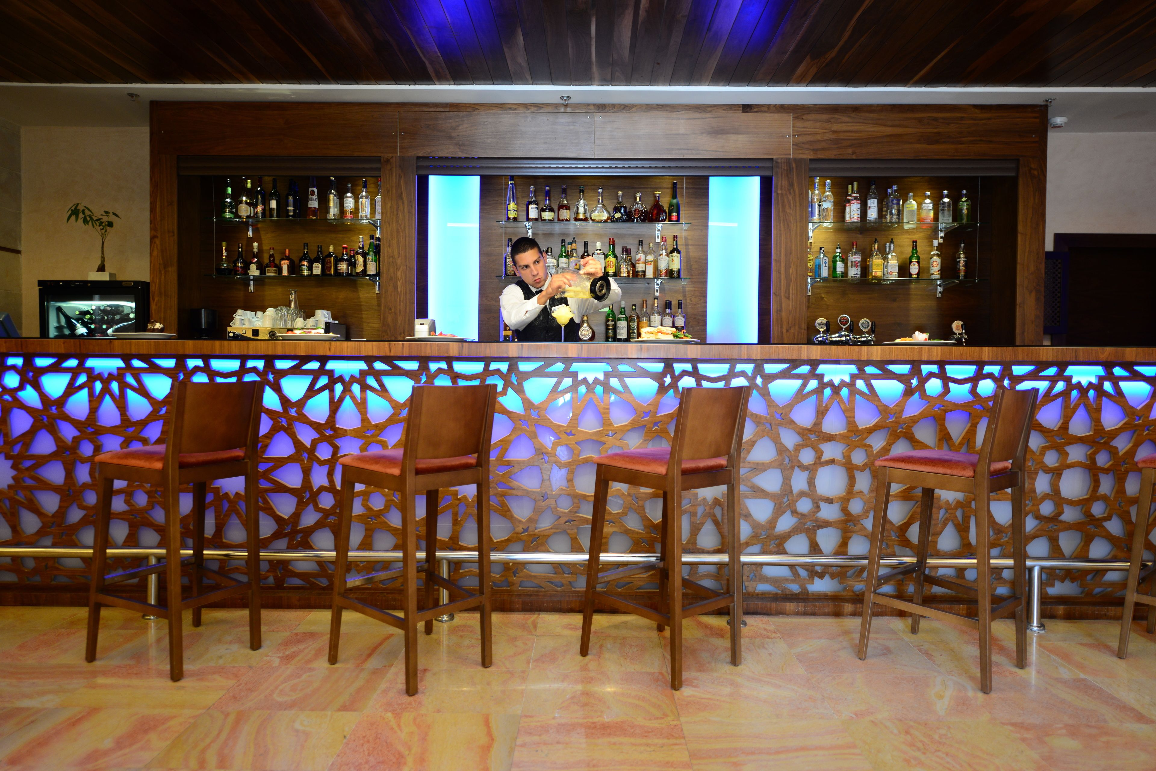 A bar area with wooden chairs, a bartender preparing drinks, and shelves stocked with bottles.