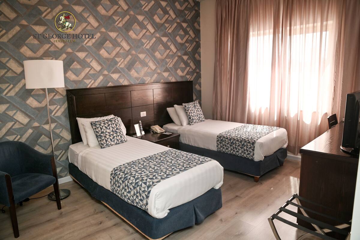 A hotel room with two beds, a desk, a chair, and a lamp.