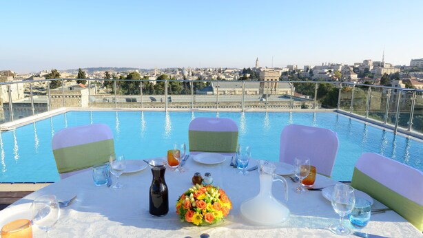 Mesa de comedor dispuesta con sillas, un jarrón de flores y vista a un paisaje urbano sobre una piscina.