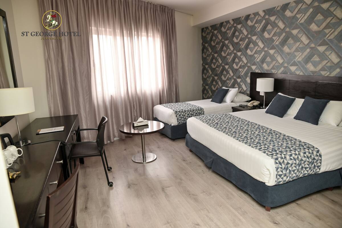 A hotel room with two beds, a desk, a chair, a small table, and a patterned wall.