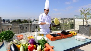 Un chef preparando comida en un balcón con vistas a la ciudad, verduras frescas y equipo de cocina.