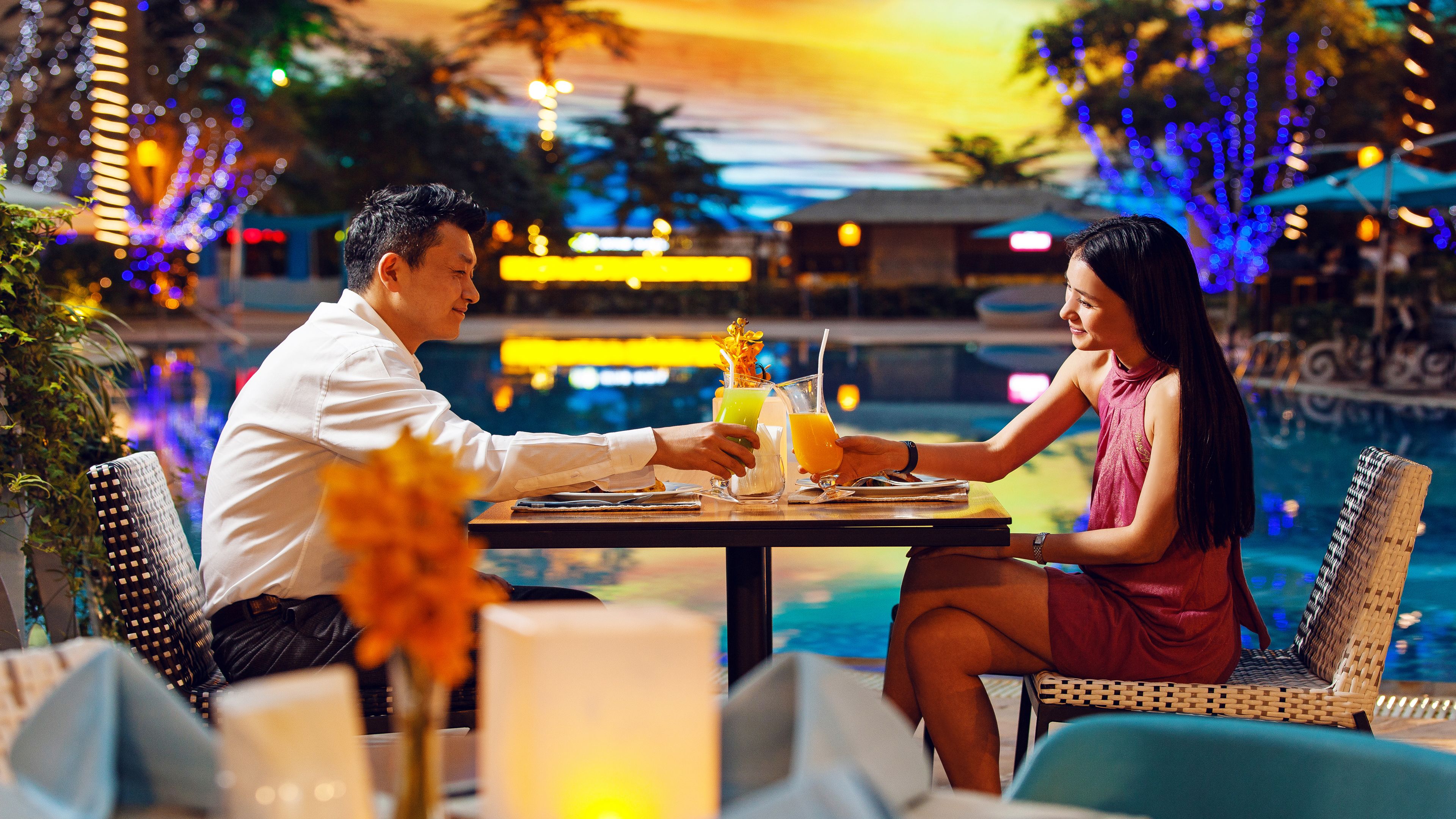 A dining table with two people seated, a poolside setting, and decorative lights in the background.