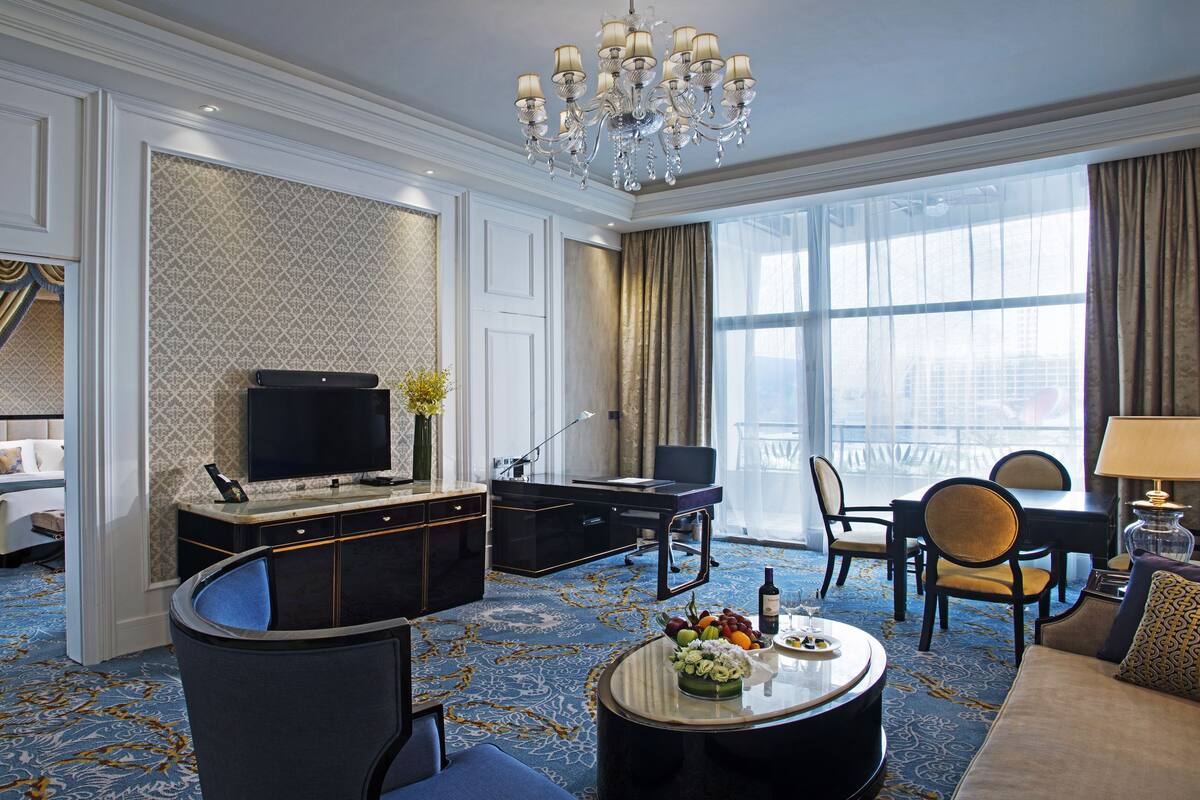 A hotel room with a large bed, a desk, a television, a dining area, and a chandelier.