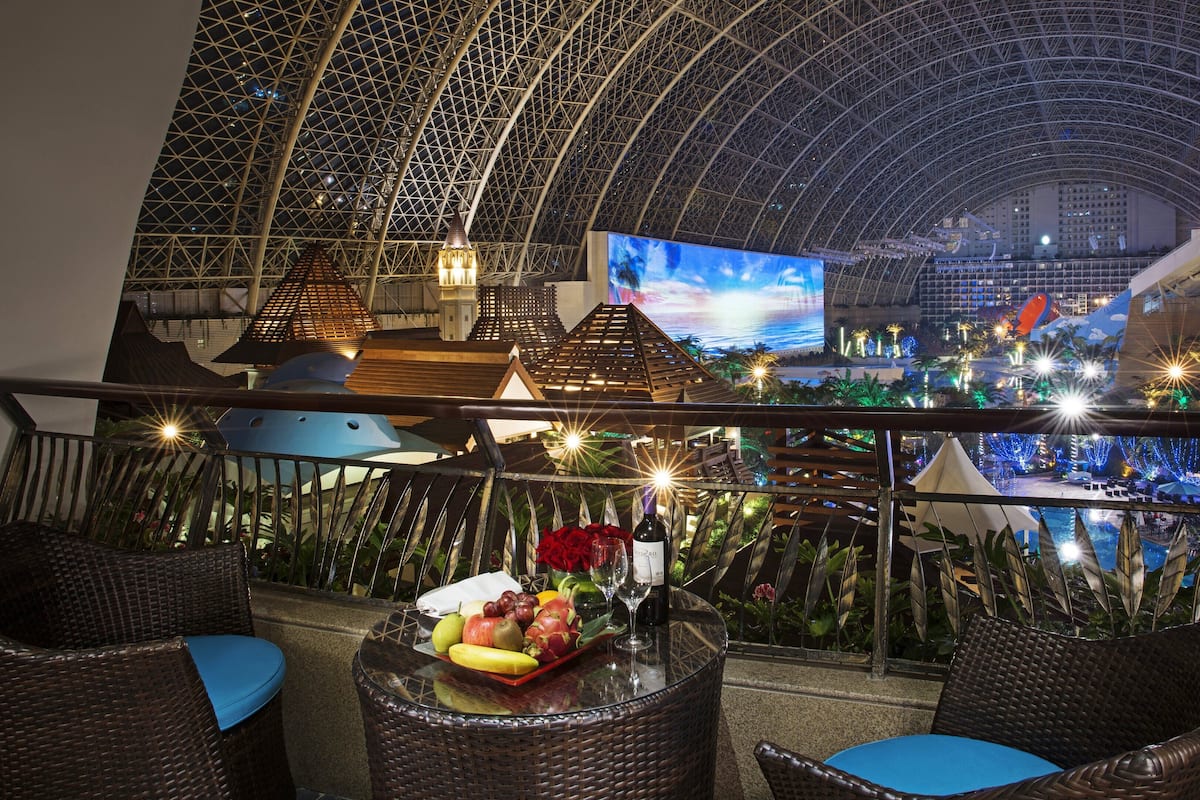 A rooftop bar with wicker chairs, a table with fruit and a bottle, and a large TV screen displaying a beach scene.