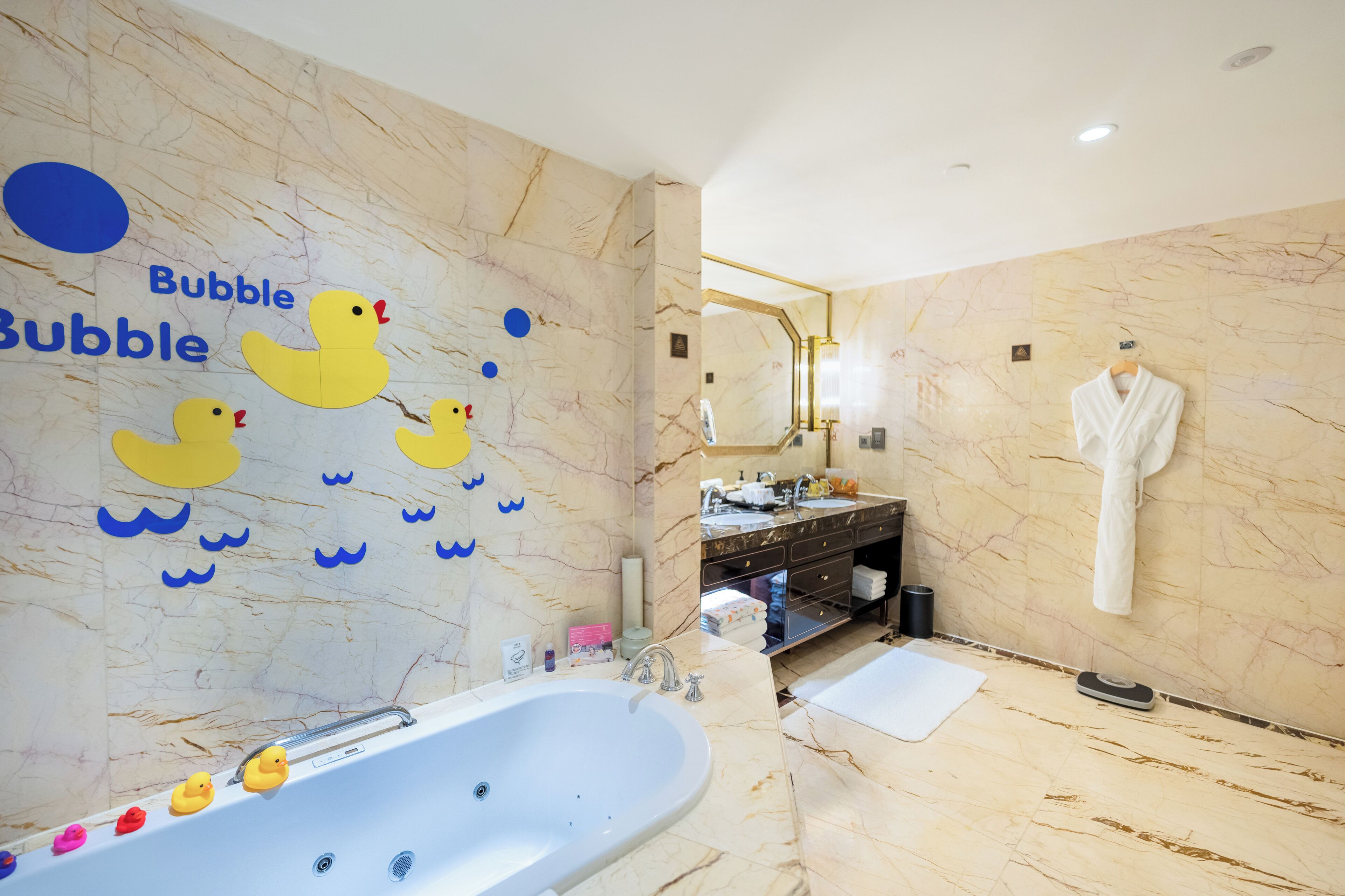 A modern bathroom with a marble floor, a large bathtub with rubber ducks, a vanity with a mirror, and a white robe hanging on the wall.