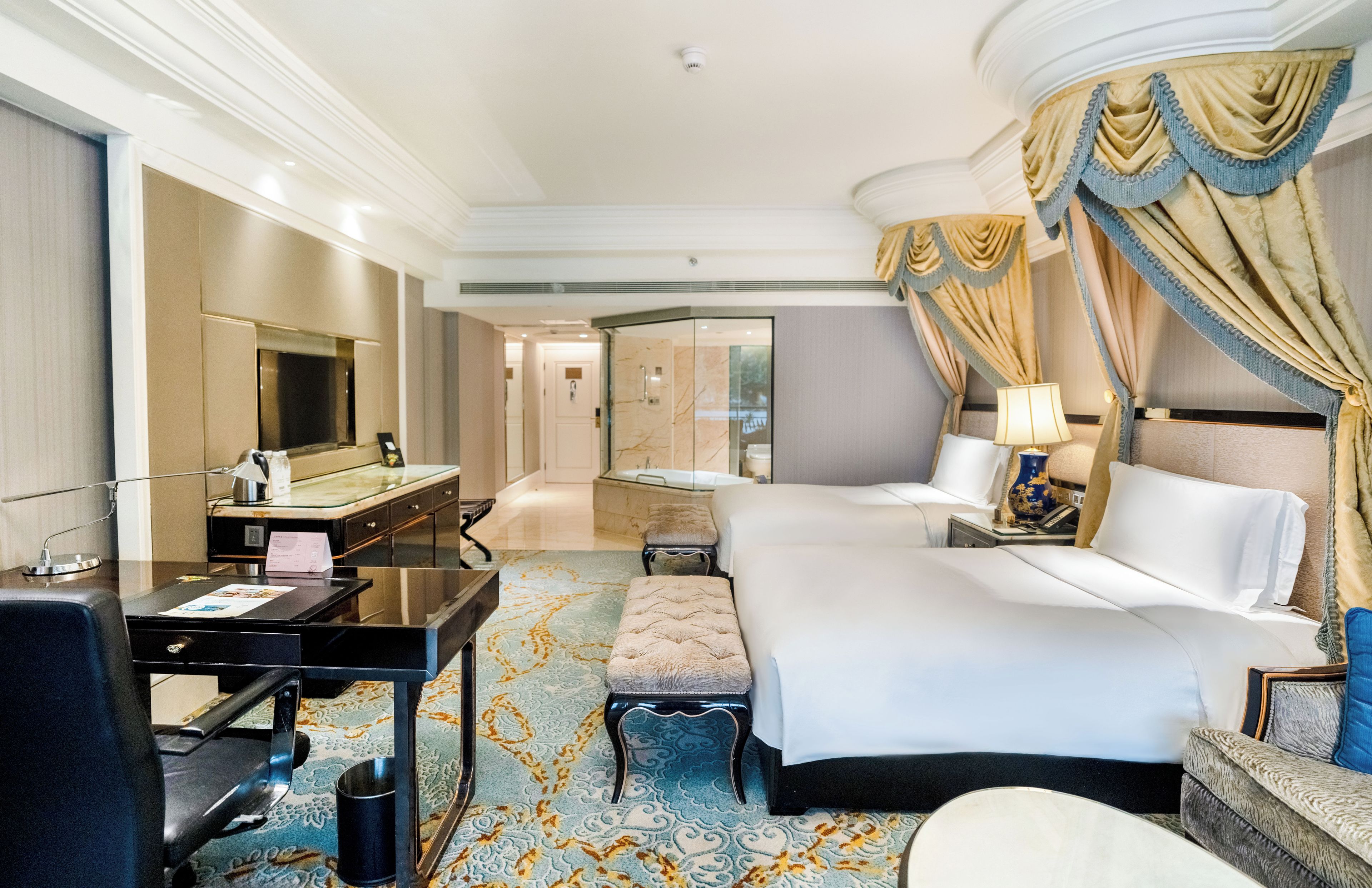 A hotel room with a bed, a desk, a chair, a television, and a bathroom visible through a glass door.