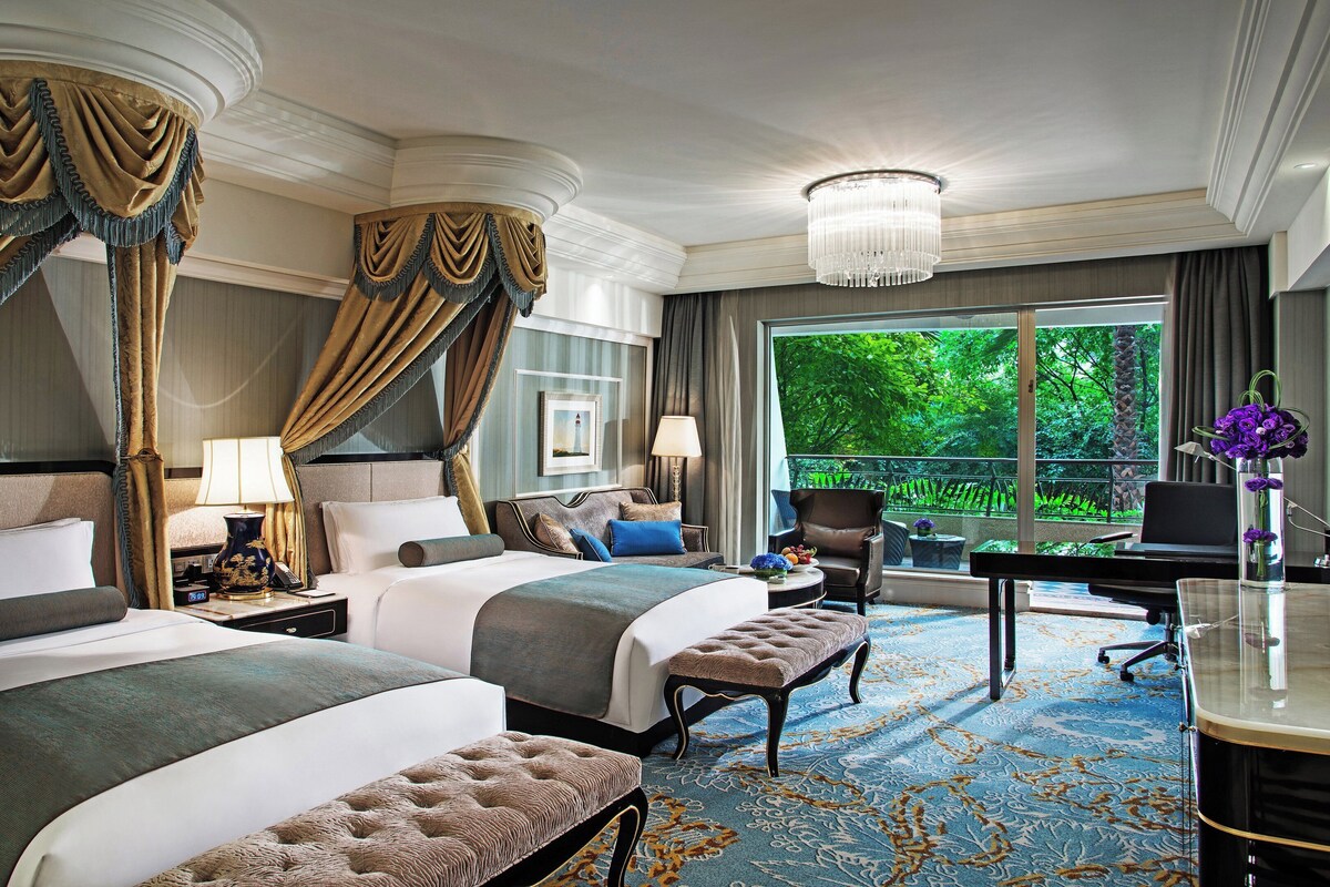 A hotel room with two beds, a desk, a chair, a computer, and a view of greenery.