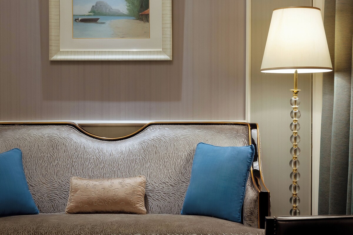 Habitación de hotel con un sofá cama con textura, un cuadro enmarcado en la pared, una lámpara de pie y un sillón de cuero oscuro.