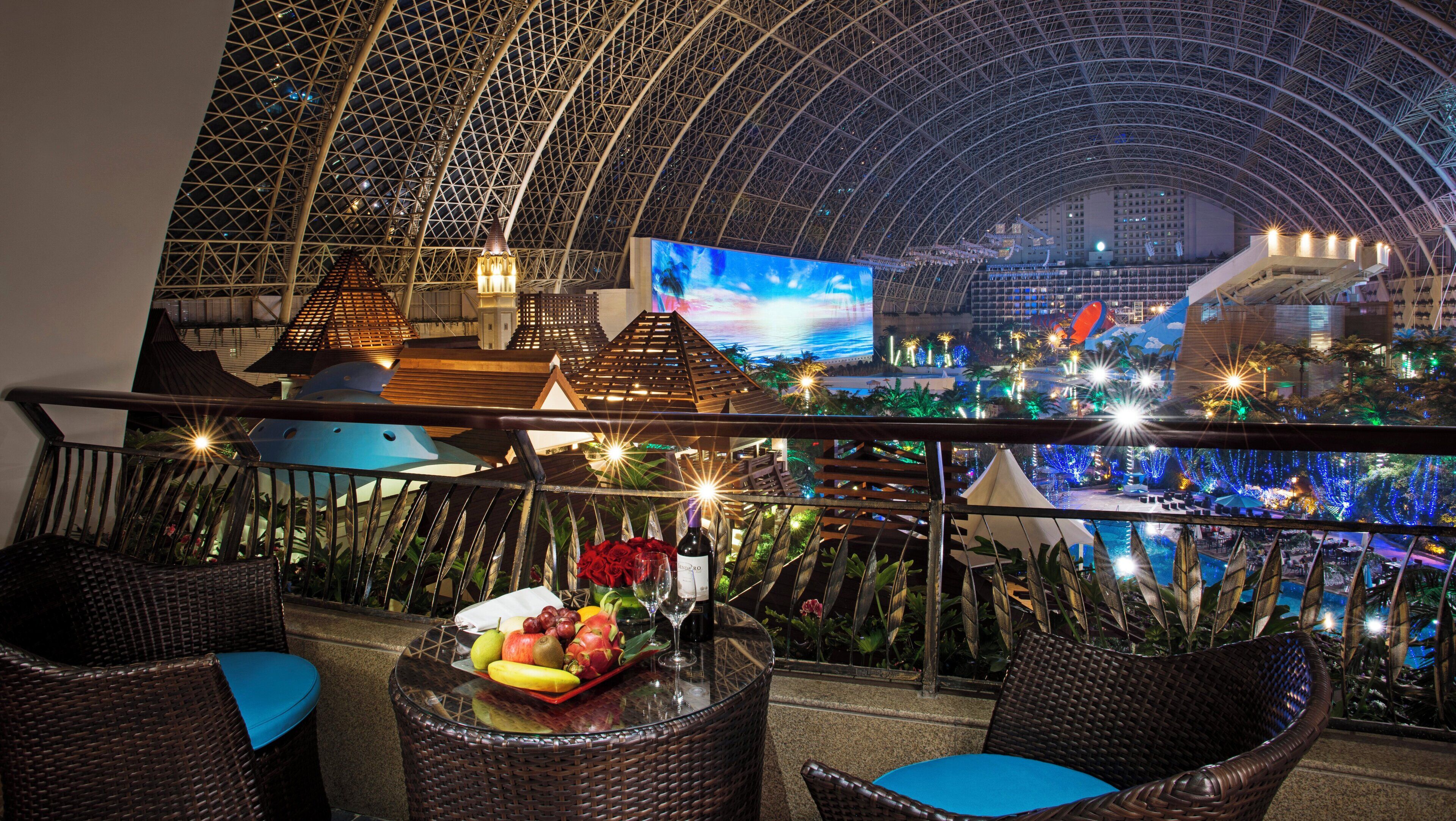 A rooftop bar with wicker chairs, a table with fruit and wine, and a view of a cityscape with a large screen.