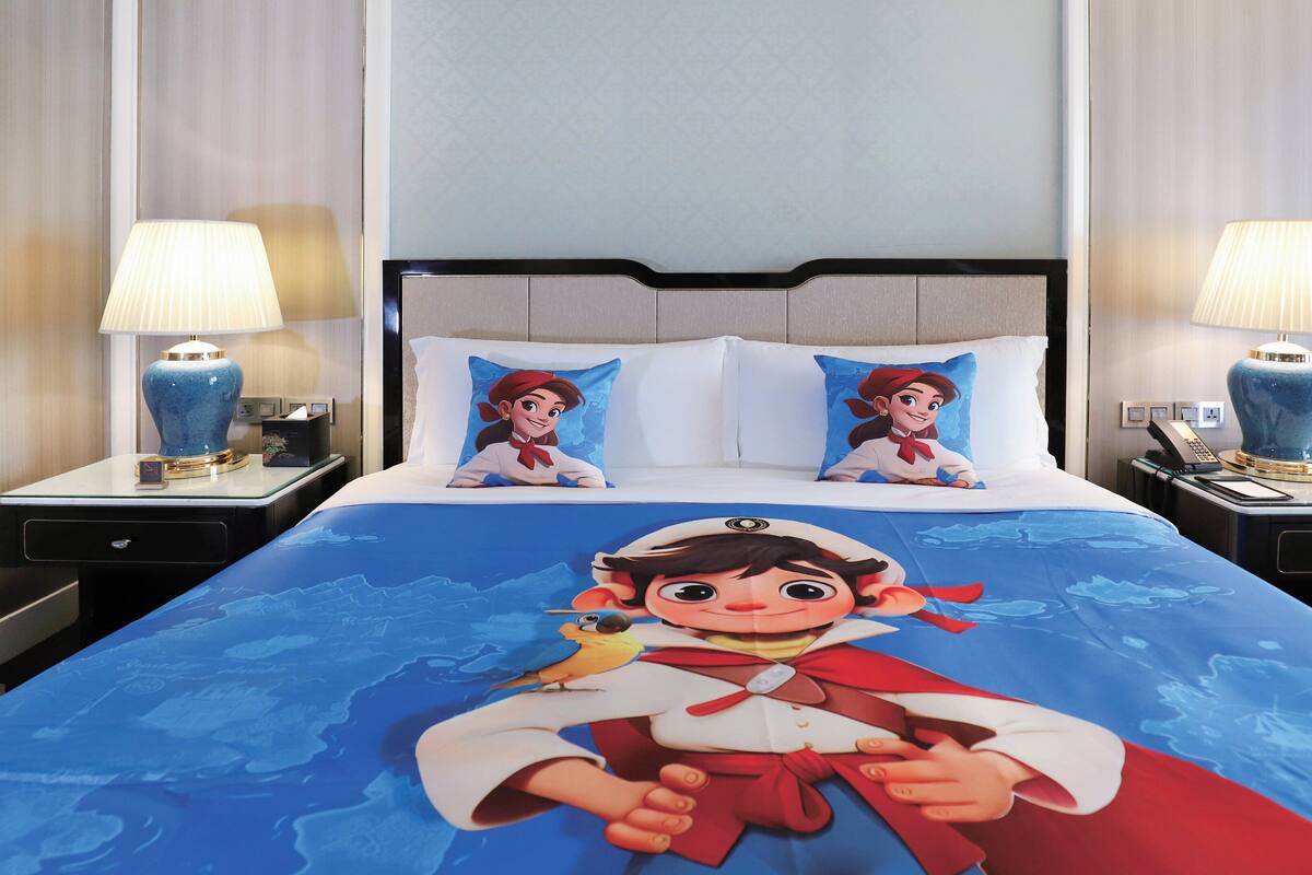 A hotel room with a large bed featuring a cartoon character bedspread, two bedside lamps, a nightstand with a phone, and a chair.