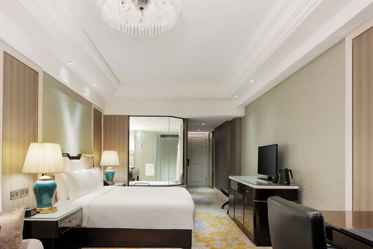 A hotel room with a large bed, a desk with a computer, a chair, a small table with fruit, and a television.