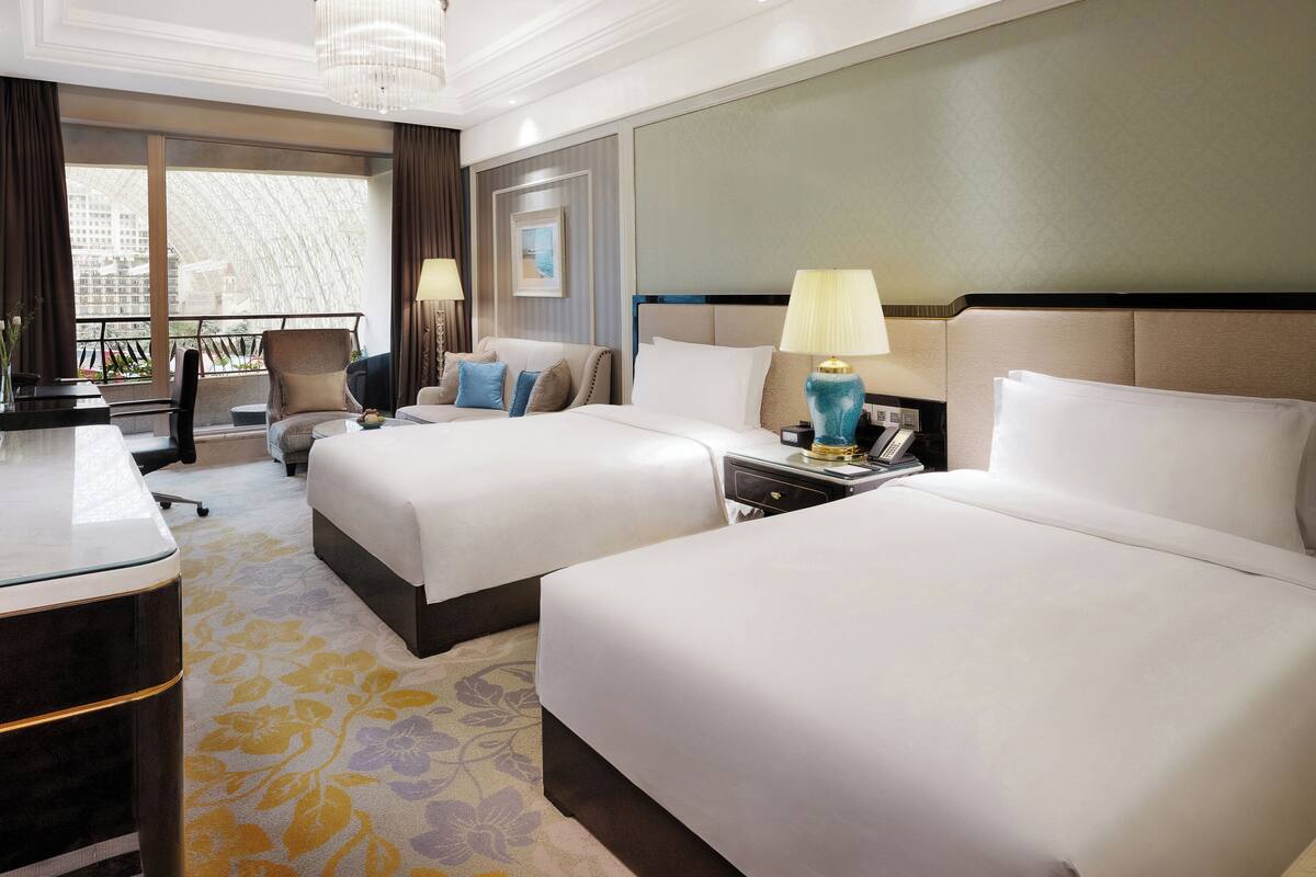 A hotel room with two beds, a desk, a chair, a lamp, and a view of a cityscape.