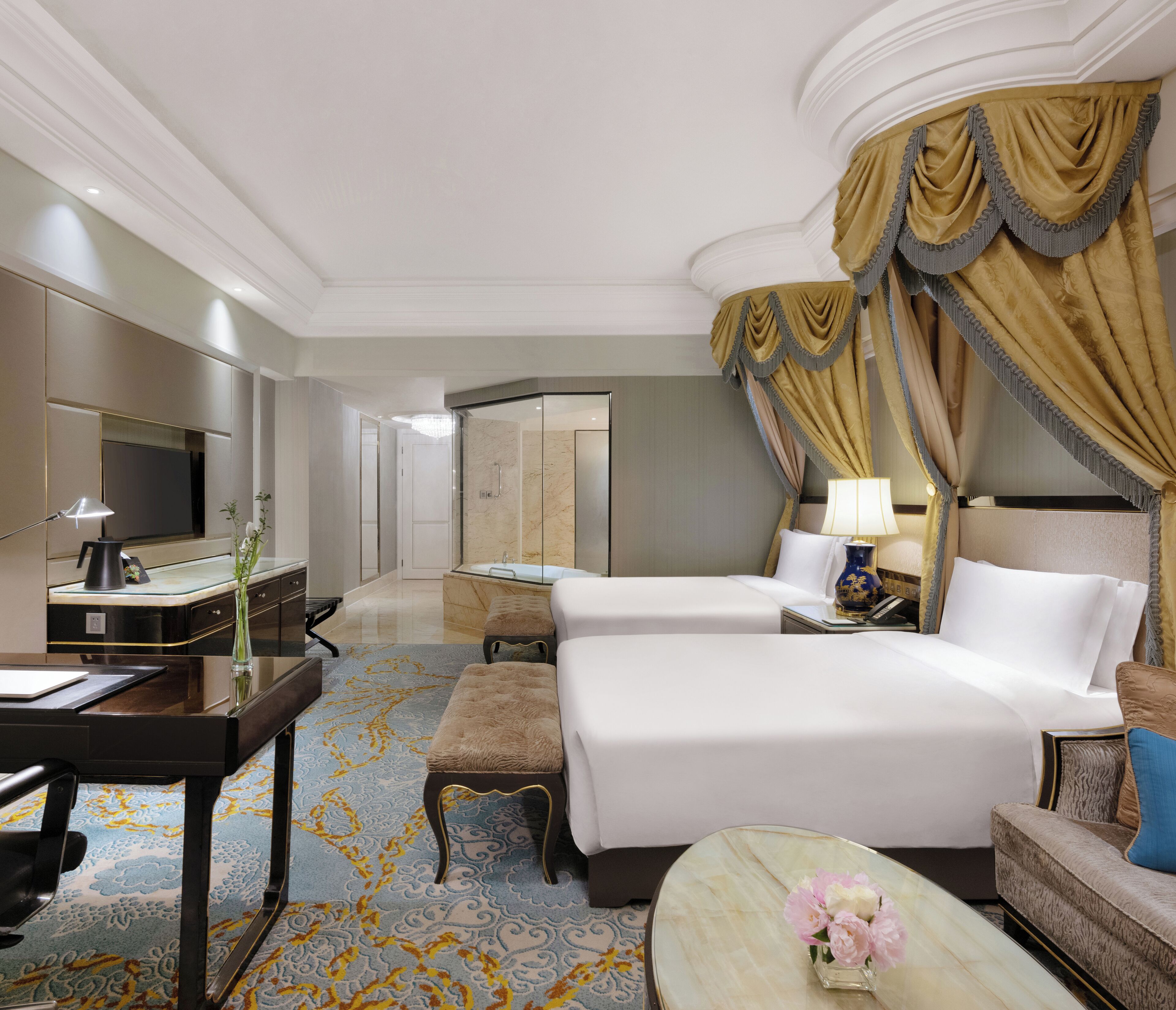 Quarto de hotel com uma cama grande, uma escrivaninha com computador, uma cadeira, uma mesinha com um vaso de flores e zona de duche.