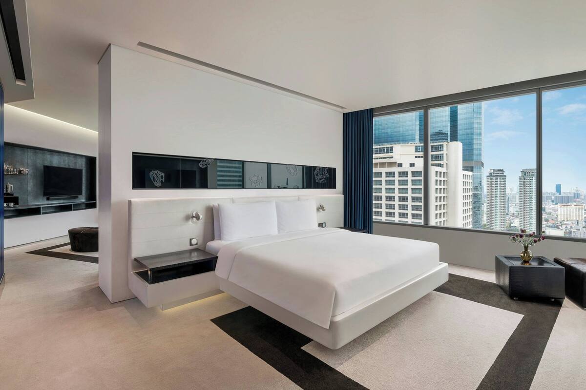 A modern hotel room with a large bed, a built-in TV, a city view, and a minimalist design.