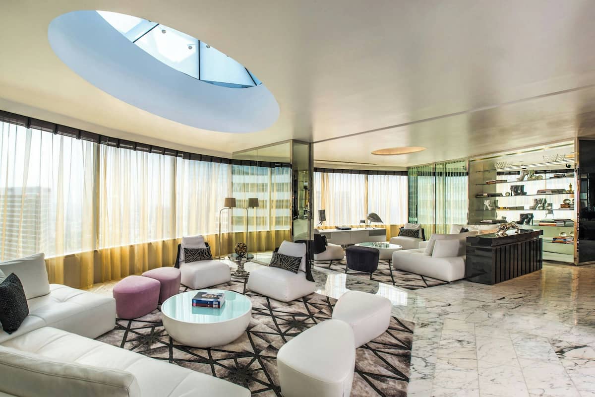 A modern living room with a high ceiling, large windows, and a central seating area.