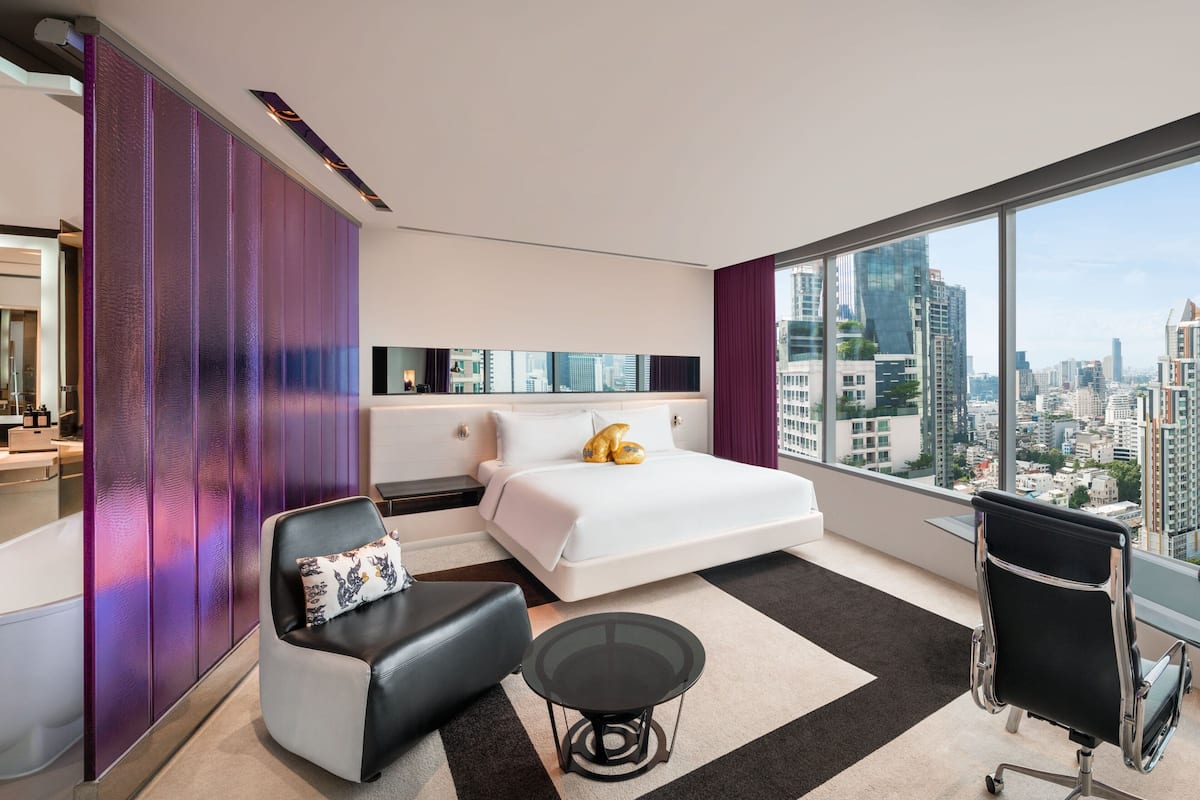 A modern hotel room with a large bed, a desk, a chair, and a view of the cityscape.