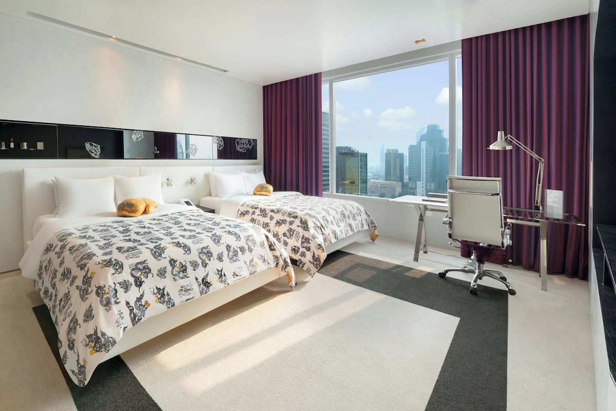 A hotel room with two beds, a desk, a chair, and a large window with a city view.