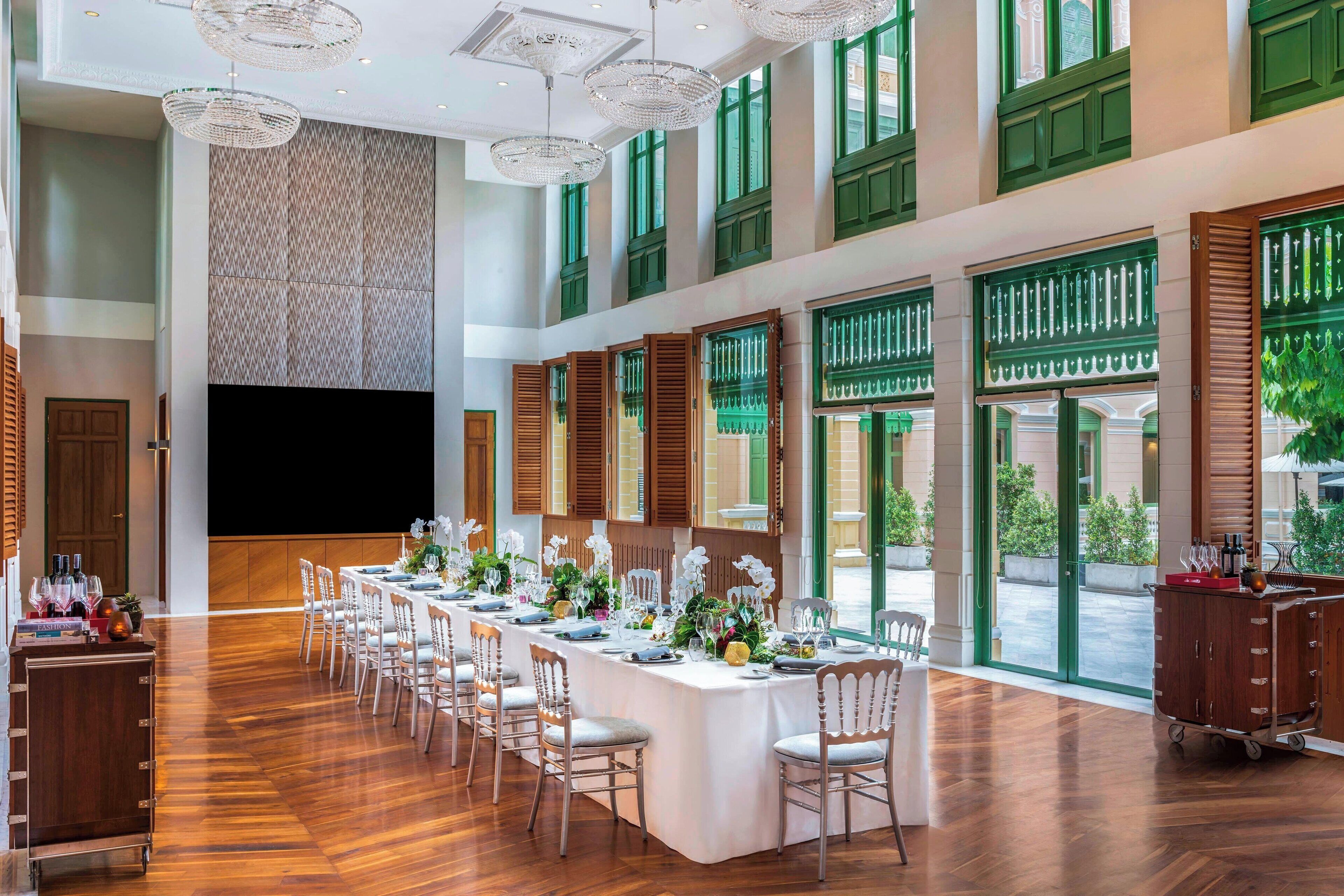 A spacious dining area with a long table set for a meal, large windows with green shutters, and a modern chandelier.