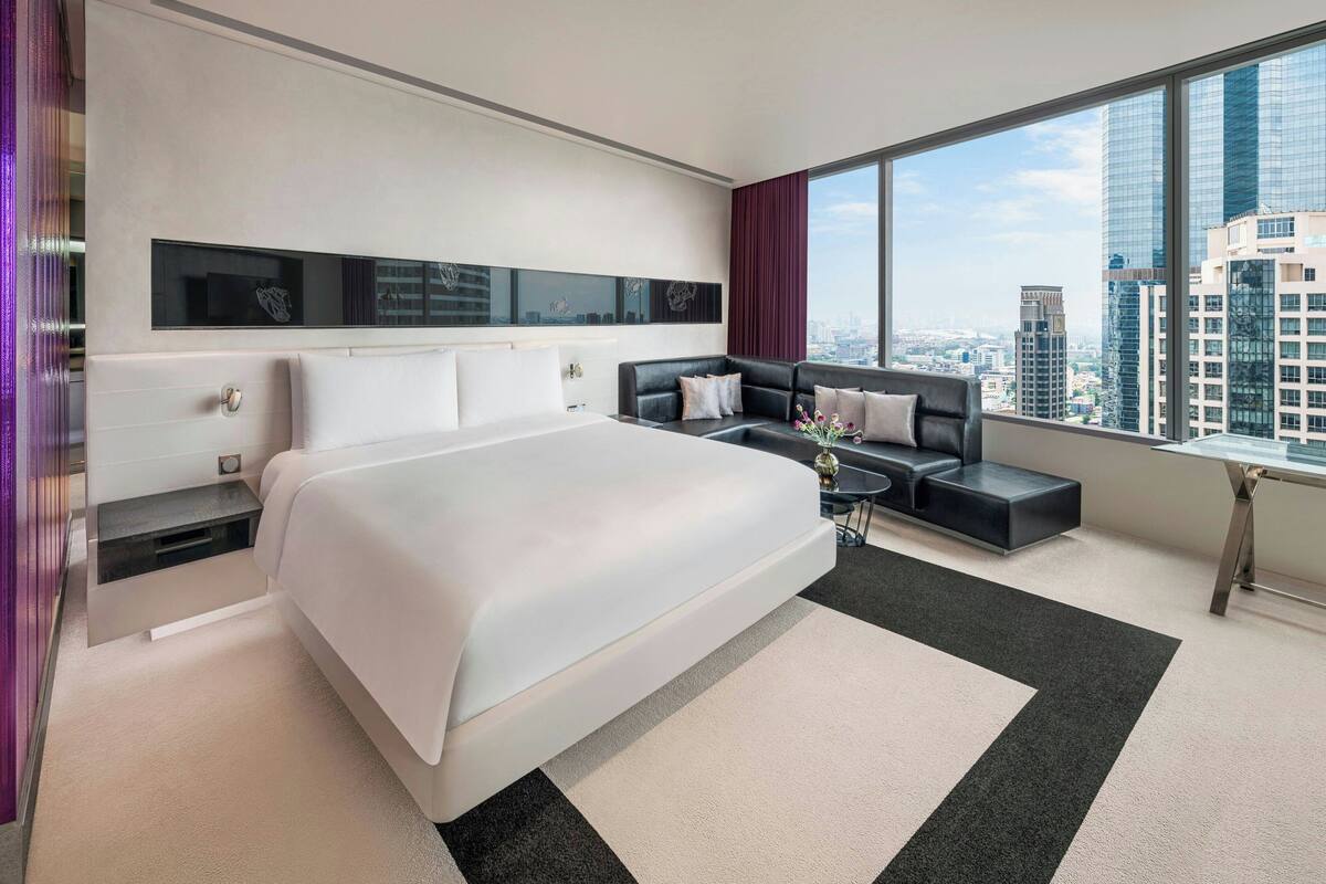 A modern hotel room with a large bed, a sofa, a small table, and a view of the cityscape through large windows.