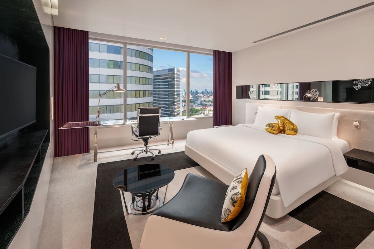 A modern hotel room with a large bed, a desk, and a chair. There is a large window with a view of buildings.