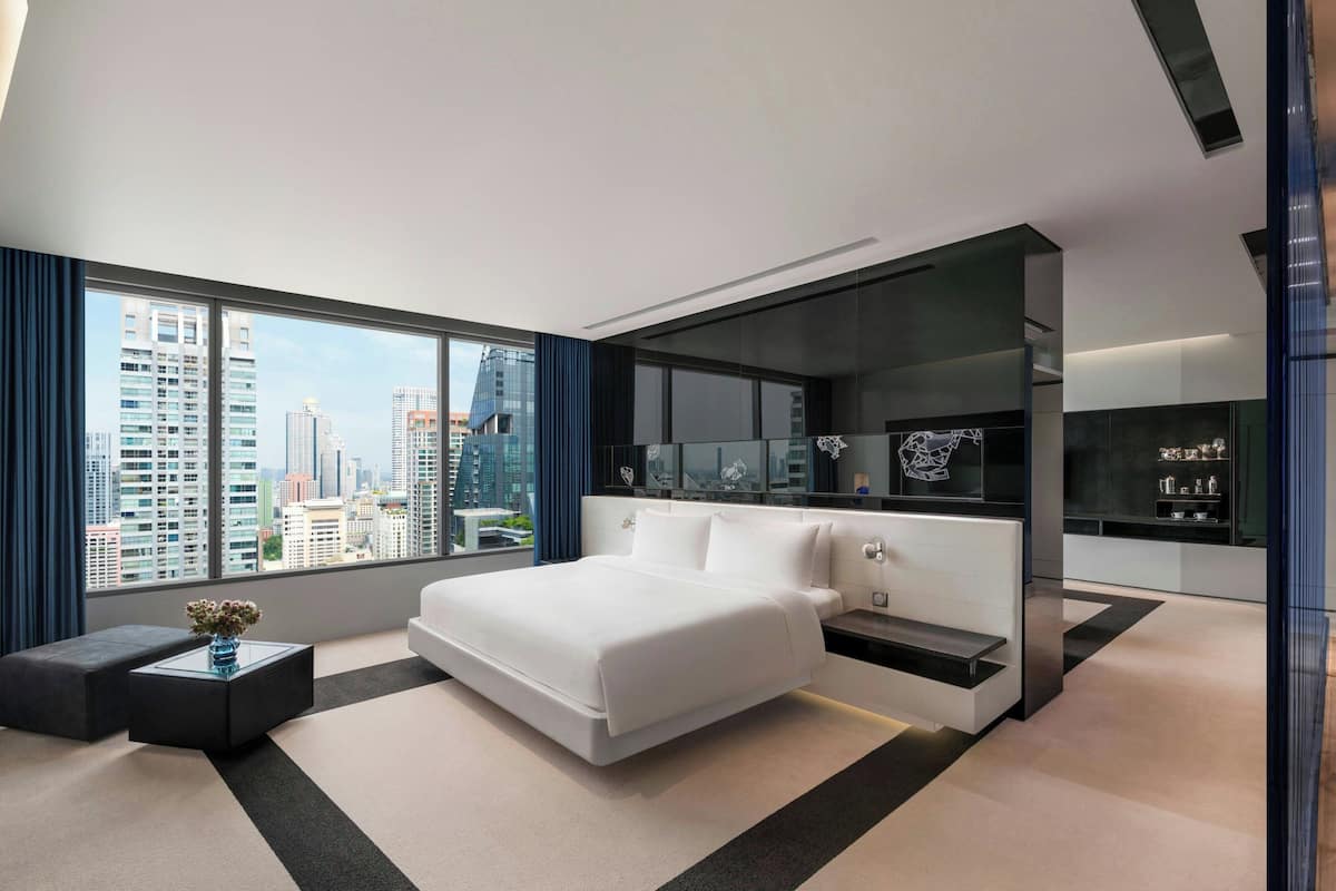 A modern hotel room with a large bed, a bedside table, a nightstand, a glass partition, and a view of the city skyline.