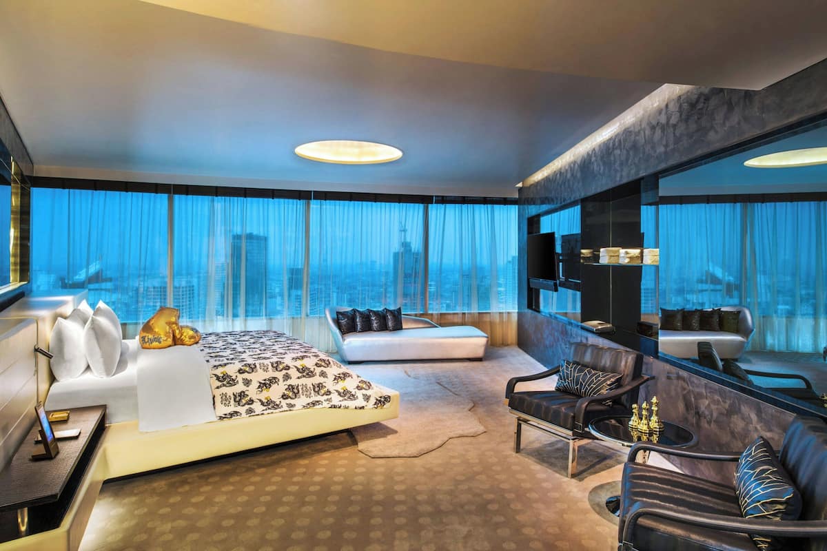 A modern hotel room with a large bed, a seating area, a desk, and a large window offering a city view.