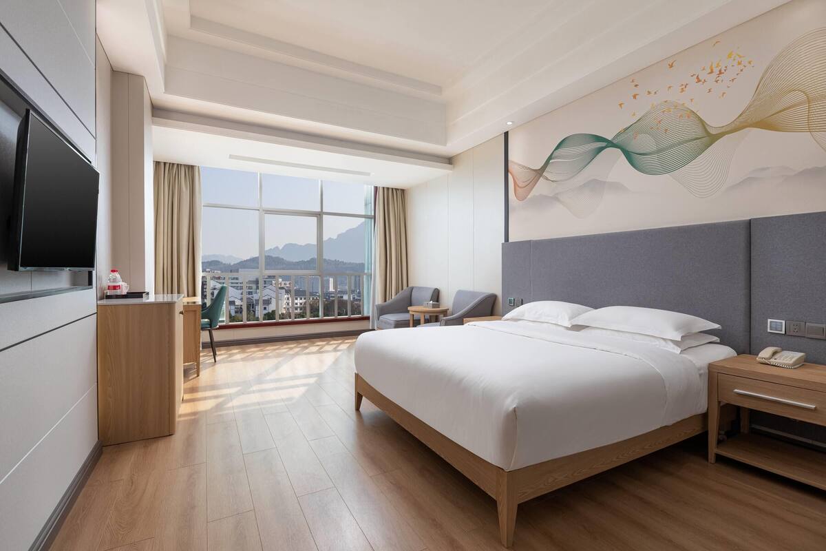 Una habitación de hotel moderna con una cama grande, televisor, escritorio y vistas a la ciudad a través de amplias ventanas.