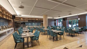 Un restaurante amplio con múltiples mesas y sillas, una zona de cocina y ventanas grandes que proporcionan luz natural.