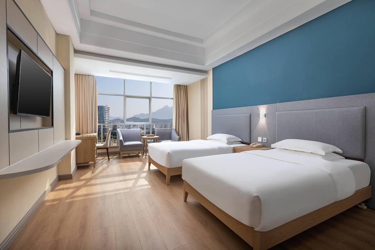 Habitación de hotel con dos camas, un escritorio, una silla, un televisor y un ventanal con vistas a la ciudad.