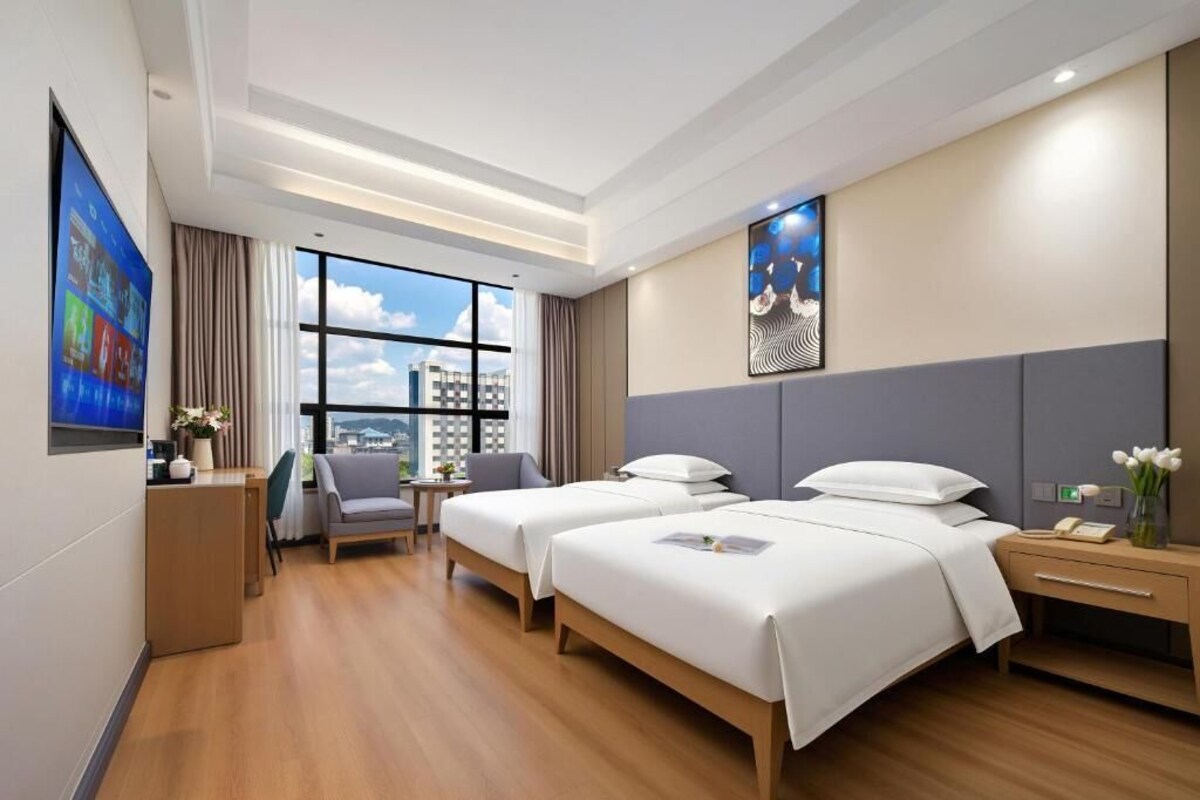 A hotel room with two beds, a desk, a chair, a small table, and a large window with a city view.