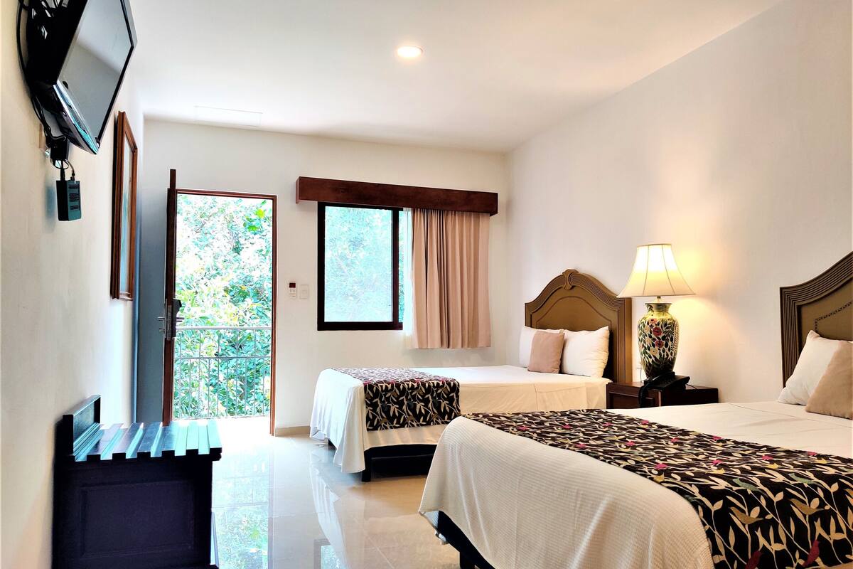 Habitación de hotel con dos camas, un televisor montado en la pared, una ventana con cortinas y una lámpara en la mesita de noche.