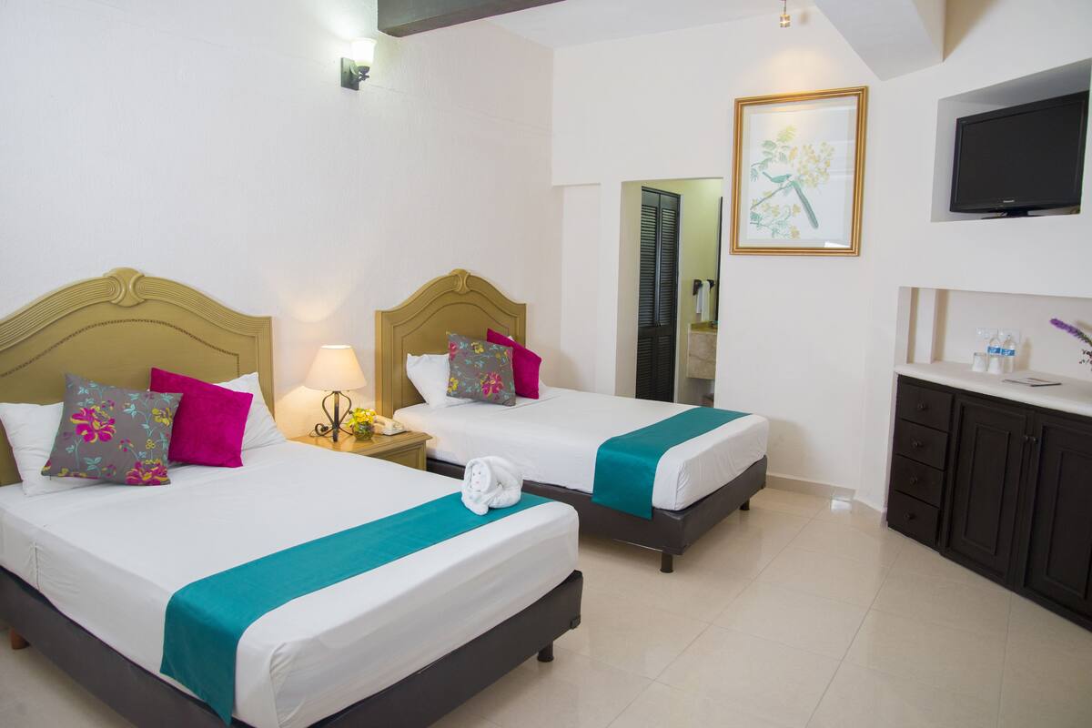 Habitación de hotel con dos camas, cada una con cubrecama color turquesa y almohadas decorativas. Se observa un mesita de noche con una lámpara y una pequeña mesa con un florero. Un cuadro enmarcado cuelga en la pared, y un televisor está montado sobre un mueble oscuro.