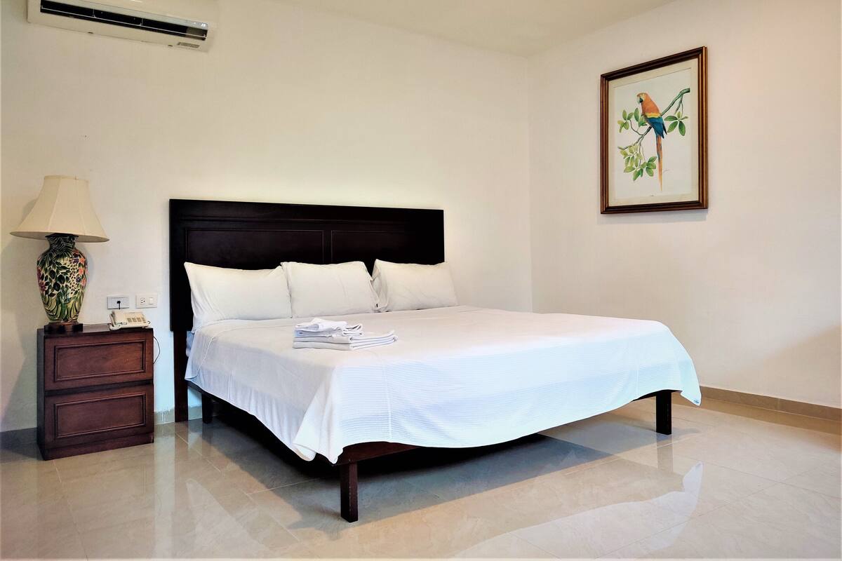 Una cama bien hecha con sábanas blancas, un cabecero oscuro, una lámpara de noche y un cuadro de un pájaro en la pared.