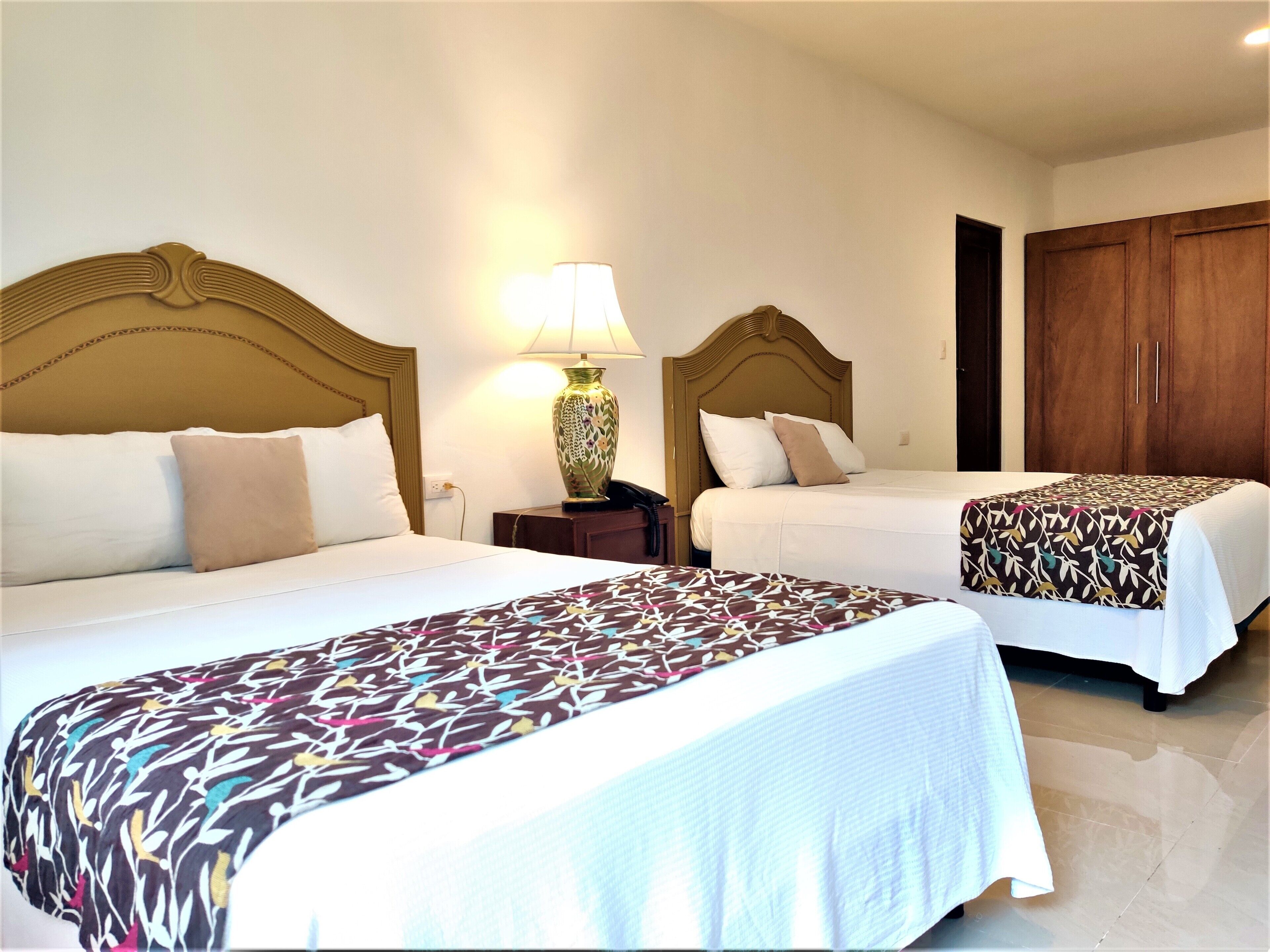 A hotel room with two beds, each with a wooden headboard, a bedside table with a lamp, and a large wardrobe.