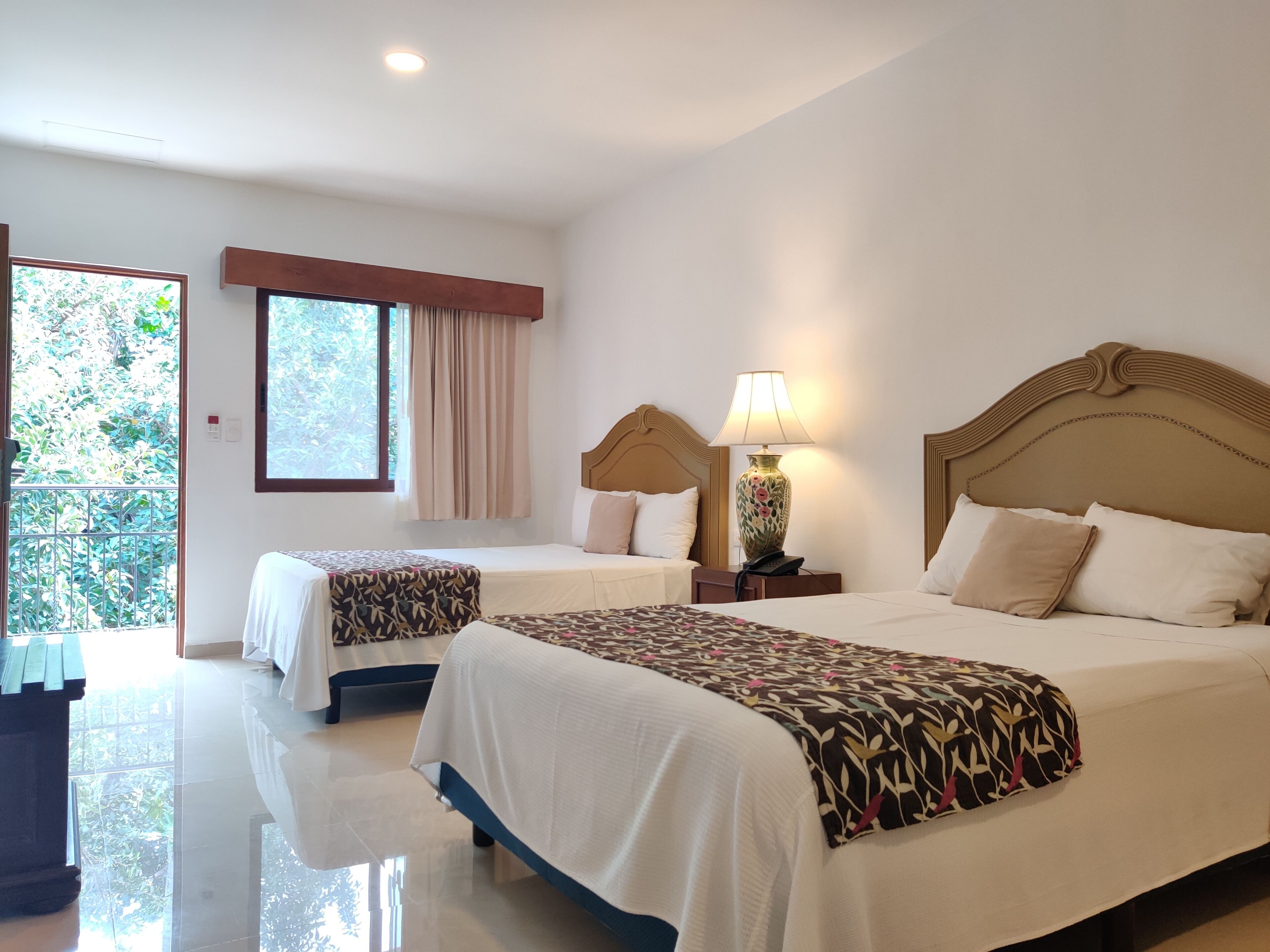 A hotel room with two beds, a large window with a view of greenery, a wooden headboard, and a bedside lamp.