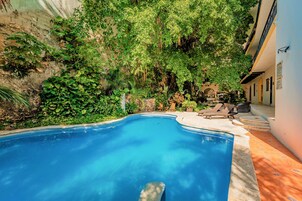 Una piscina rodeada de abundante vegetación, con un patio y sillones de descanso.