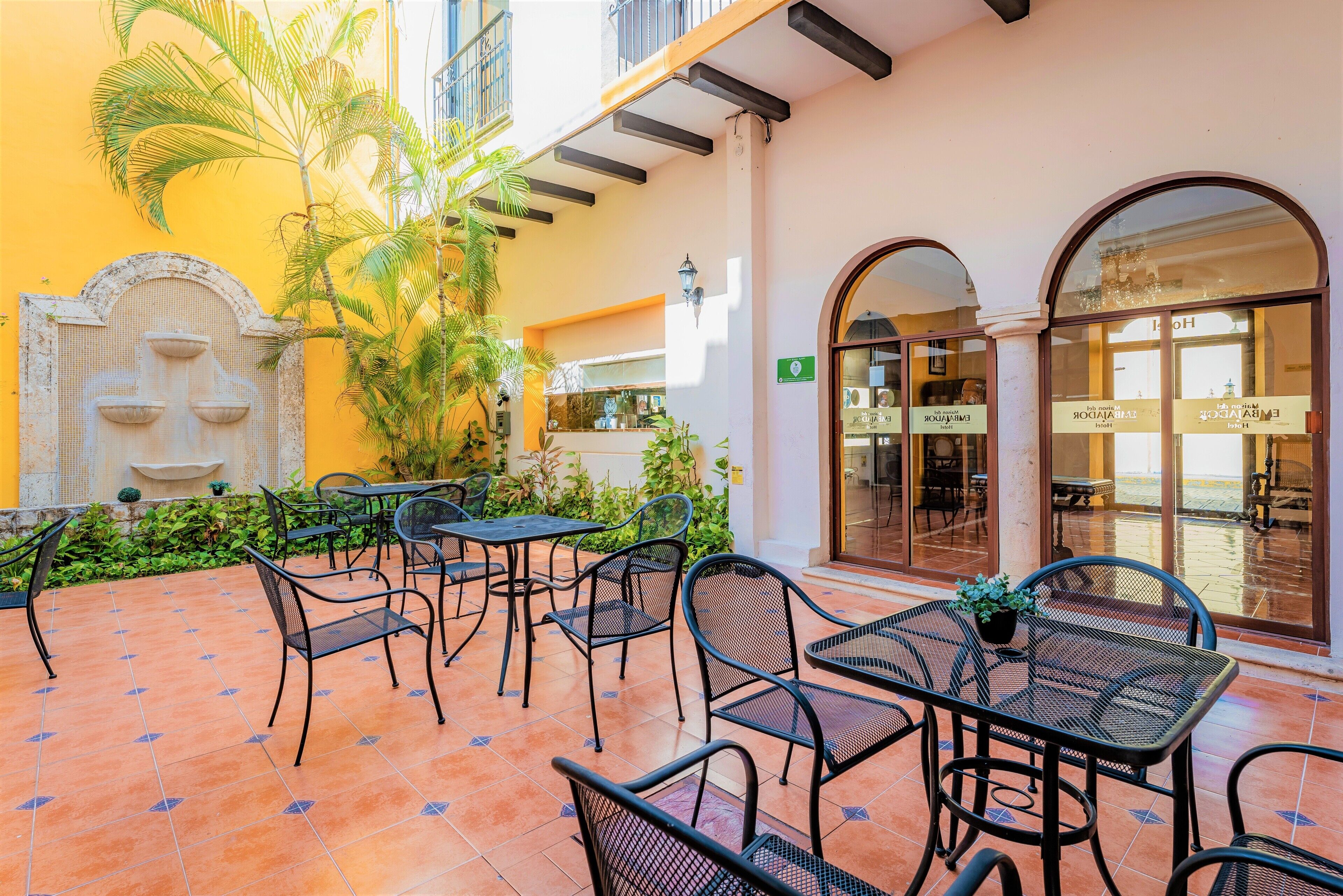 An outdoor patio with metal tables and chairs, a fountain, and a building with a yellow facade and arched windows.