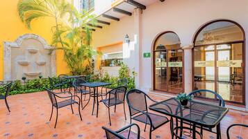 Un patio al aire libre con mesas y sillas de metal, una fuente y un edificio con fachada amarilla y ventanas arqueadas.
