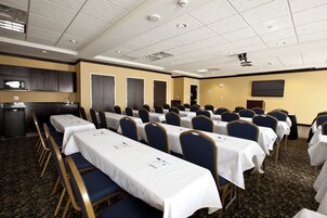 A conference room with rows of tables and chairs, a kitchenette, and a flat-screen TV on the wall.