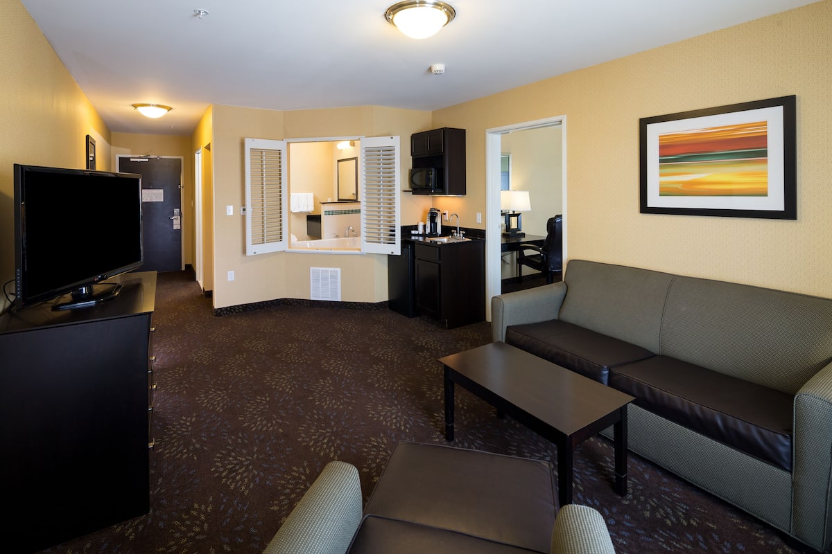 A hotel room with a flat-screen TV, a sofa, a coffee table, a dining area with a microwave, and a painting on the wall.