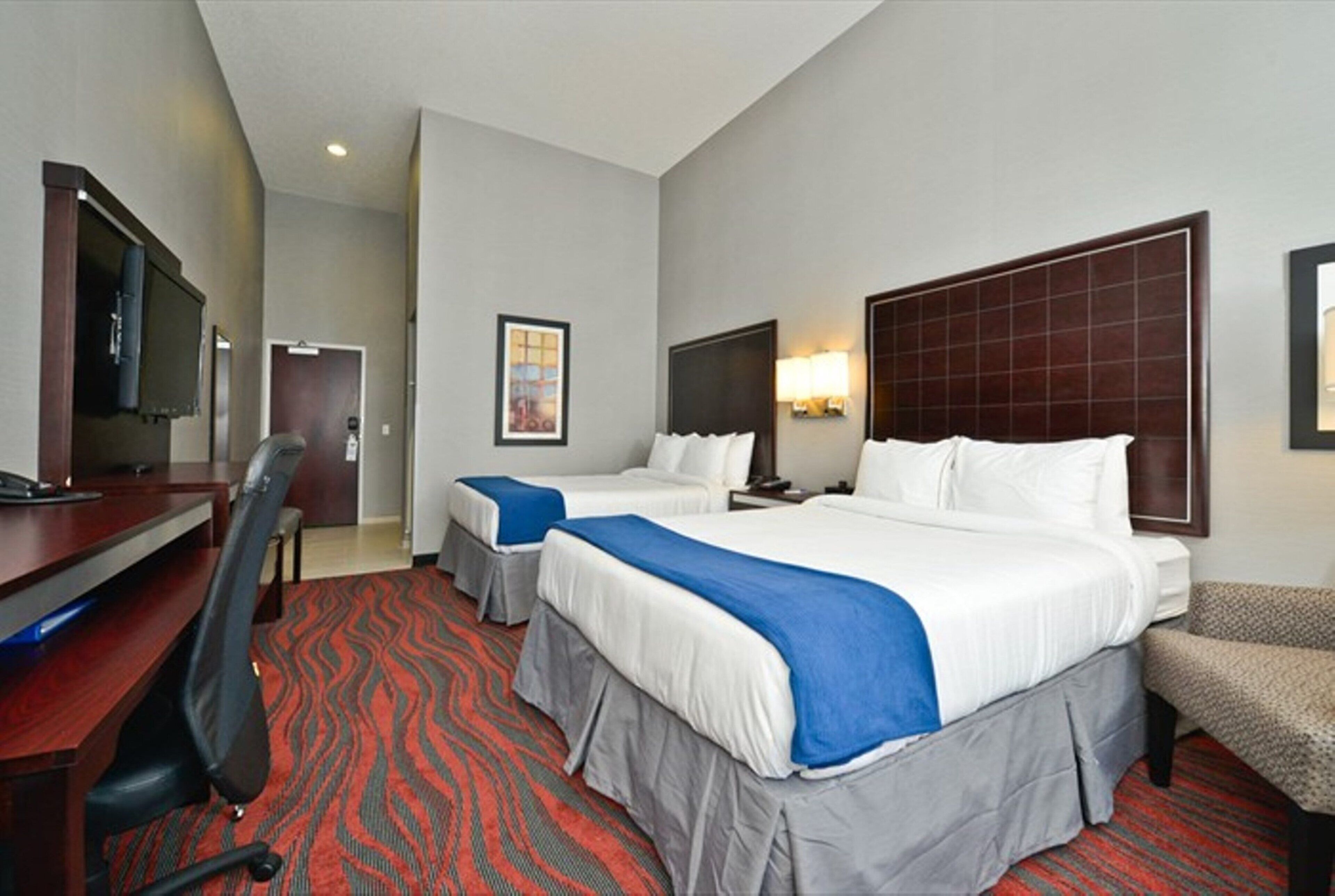 A hotel room with two beds, a desk, a chair, a TV, and a red carpet.