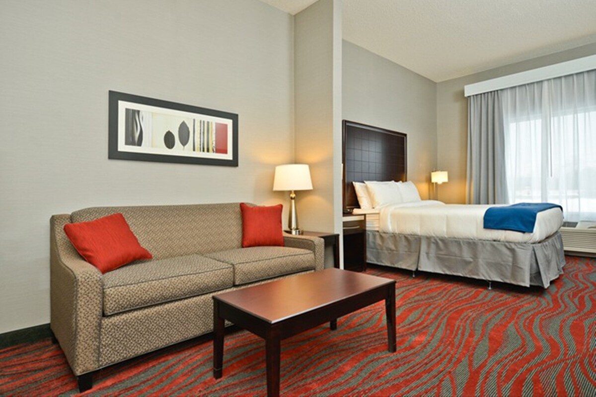 A hotel room with a sofa, a bed, a nightstand, a lamp, a framed picture on the wall, and a patterned carpet.