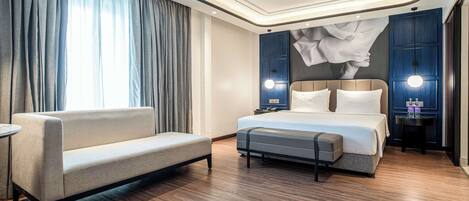 A modern hotel room with a large bed, a sofa, a small table, and a wall-mounted artwork.