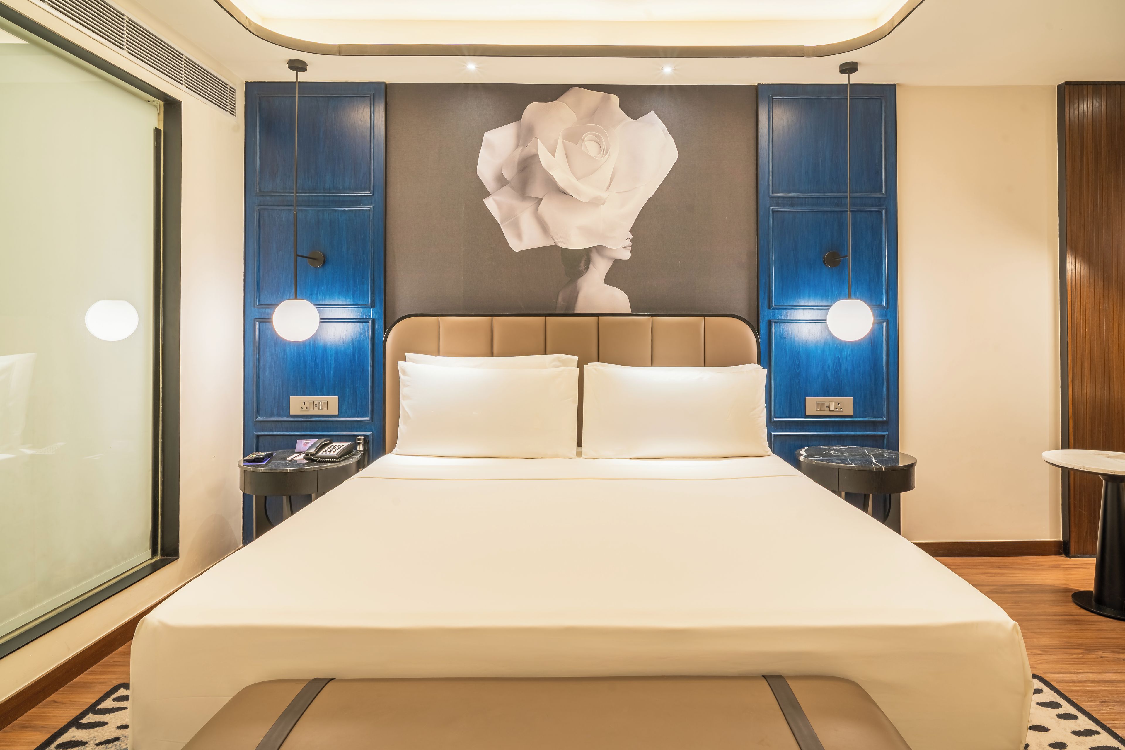 Quarto de hotel com uma cama grande, duas mesas de cabeceira, luminária fixada na parede e uma grande obra de arte abstrata.
