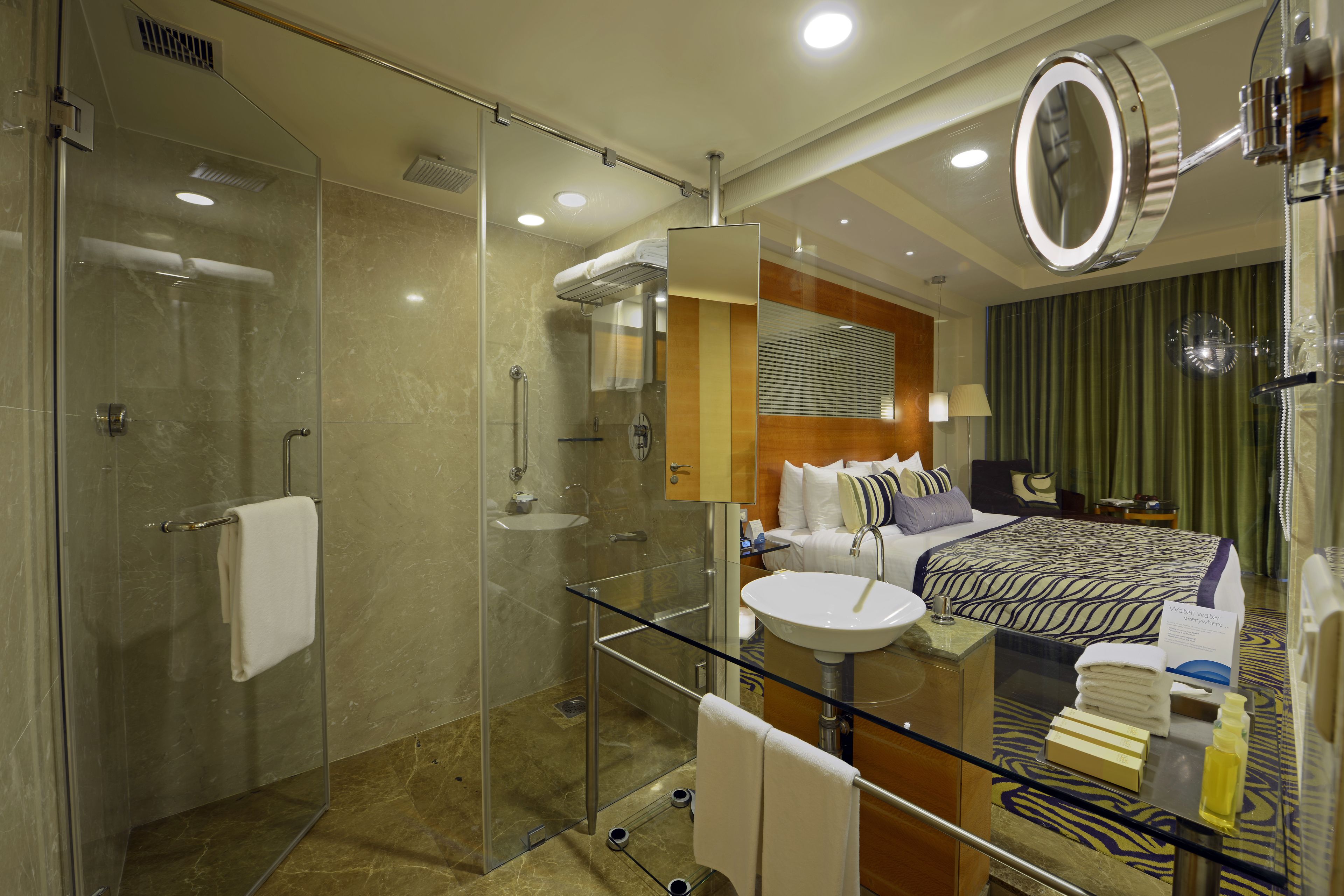 A hotel room with a glass shower enclosure, a bed with a patterned bedspread, a round mirror, and a bedside table with toiletries.